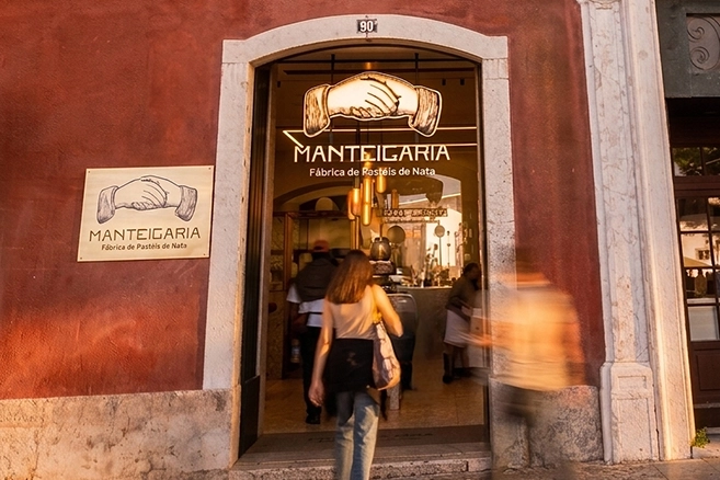 Entrada da Manteigaria numa fachada vermelha ao entardecer, com uma pessoa a caminhar